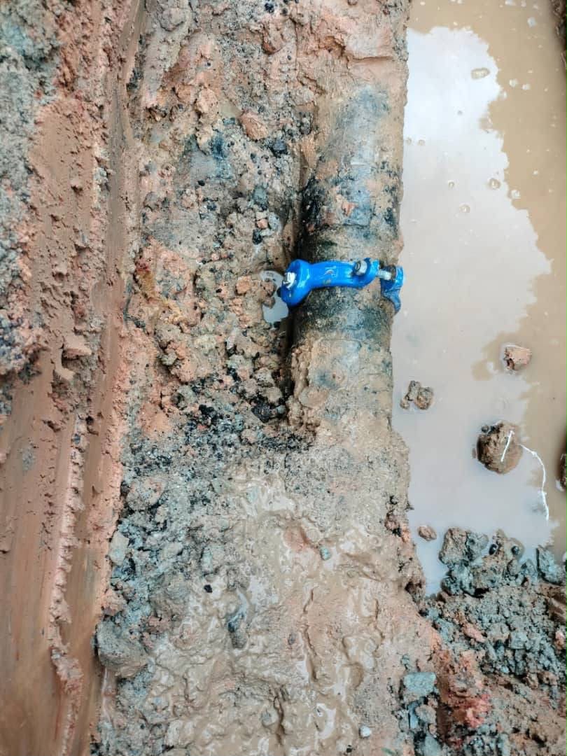 A repair clamp fitted around a corroded underground pipe at the bottom of a trench of muddy water