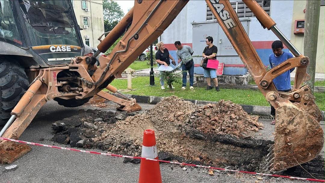A backhoe loader digging a trench through the asphalt of a residential street, watched from the verge