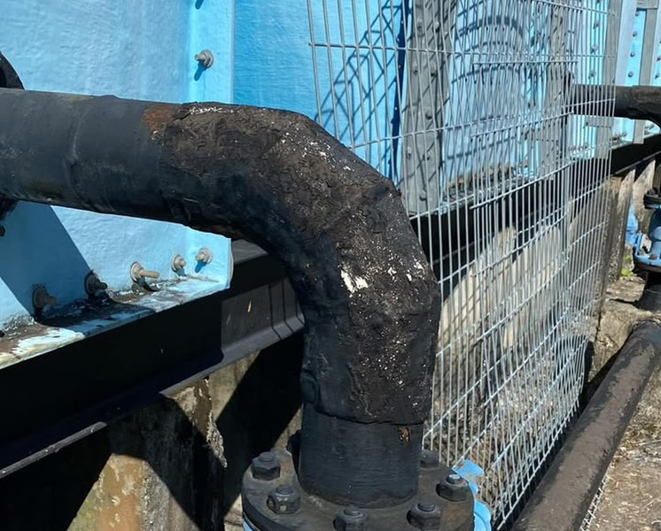 Bitumen-coated water main running into a blue gate valve against a tank wall